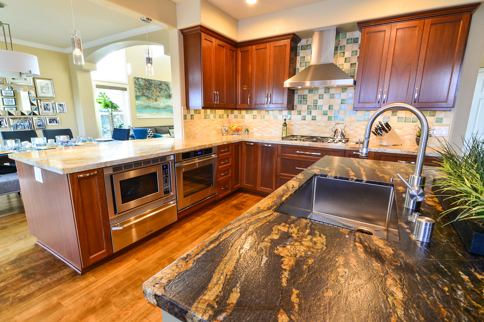 Photo of a large traditional u-shaped open plan kitchen in Tampa with a single-bowl sink, shaker cabinets, medium wood cabinets, granite worktops, green splashback, glass tiled splashback, stainless steel appliances, light hardwood flooring and an island.