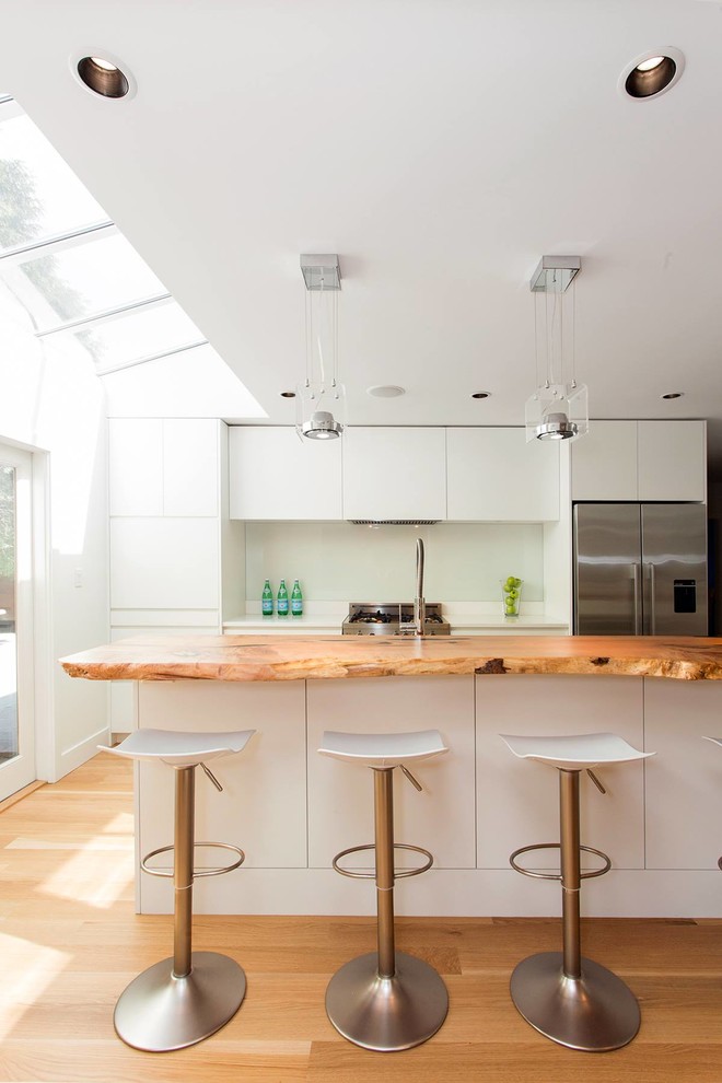 На фото: параллельная кухня в современном стиле с плоскими фасадами, белыми фасадами, деревянной столешницей, белым фартуком и островом