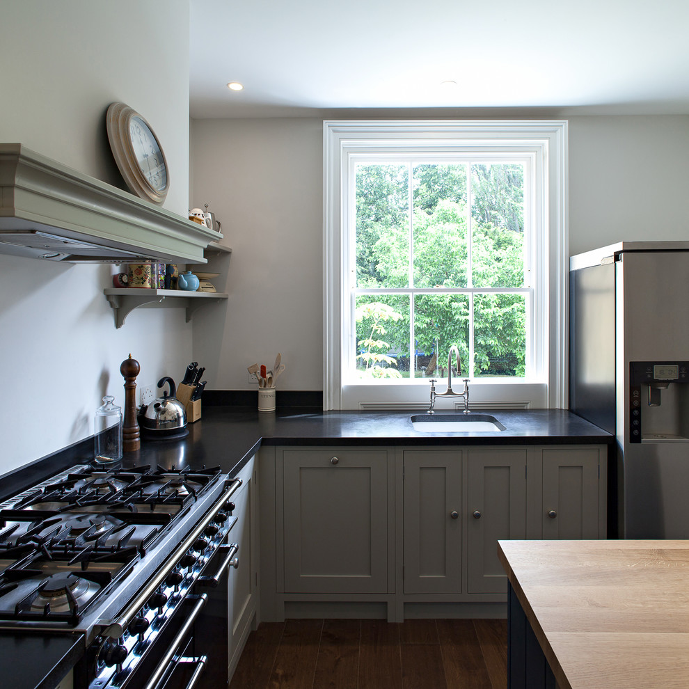Mittelgroße Klassische Wohnküche in L-Form mit Landhausspüle, Schrankfronten im Shaker-Stil, grünen Schränken, Granit-Arbeitsplatte, schwarzen Elektrogeräten, dunklem Holzboden, Kücheninsel und Küchenrückwand in Schwarz in London