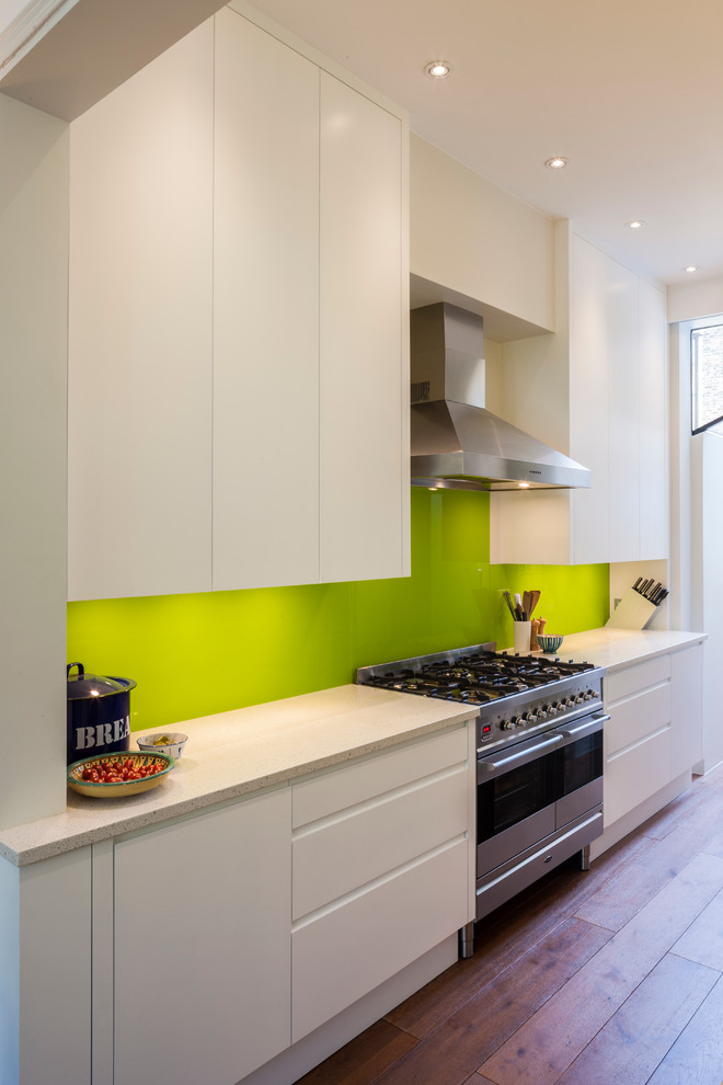 Photo of a contemporary galley kitchen in London with flat-panel cabinets, white cabinets, green splashback, glass sheet splashback, stainless steel appliances, engineered stone countertops and medium hardwood flooring.