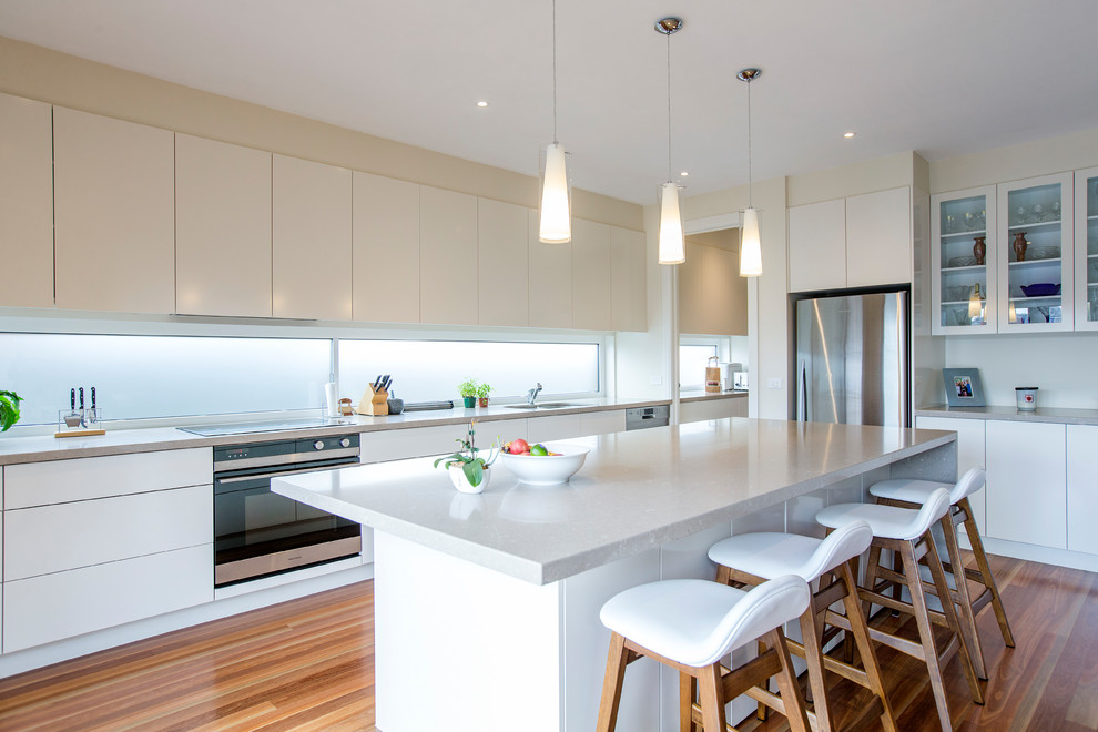 Mittelgroße Moderne Küche in L-Form mit Unterbauwaschbecken, flächenbündigen Schrankfronten, weißen Schränken, Rückwand-Fenster, Küchengeräten aus Edelstahl, braunem Holzboden, Kücheninsel, braunem Boden und grauer Arbeitsplatte in Melbourne