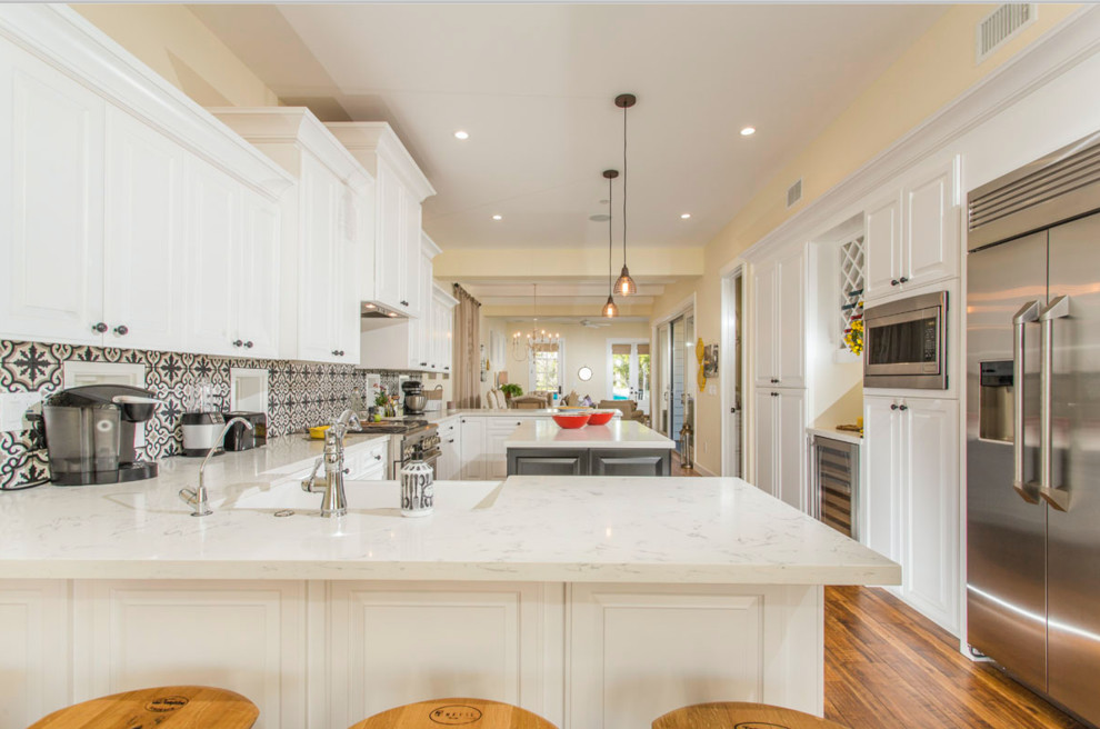 Large beach style u-shaped laminate floor and brown floor eat-in kitchen photo in San Diego with quartzite countertops, black backsplash, an island, a farmhouse sink, raised-panel cabinets, white cabinets, cement tile backsplash and stainless steel appliances