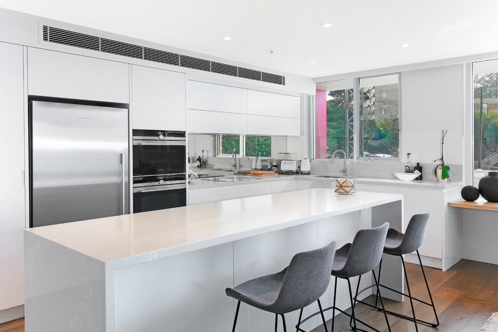 Foto di un cucina con isola centrale contemporaneo con ante bianche, paraspruzzi a specchio, elettrodomestici in acciaio inossidabile, pavimento marrone e top bianco