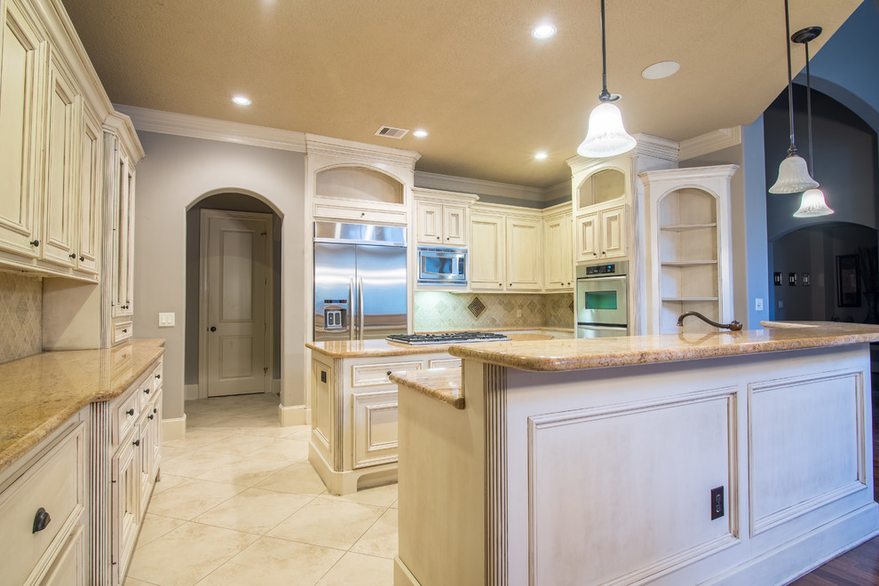 Example of a huge classic travertine floor kitchen pantry design in Houston with a double-bowl sink, raised-panel cabinets, distressed cabinets, granite countertops, beige backsplash, stone tile backsplash, stainless steel appliances and an island