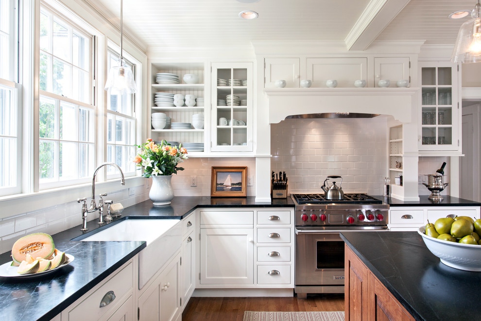 Traditional l-shaped kitchen/diner in Boston with a belfast sink, glass-front cabinets, white splashback, metro tiled splashback, stainless steel appliances, an island, soapstone worktops and medium hardwood flooring.