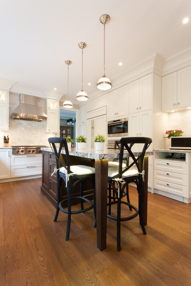 Example of a large classic u-shaped medium tone wood floor eat-in kitchen design in Vancouver with a farmhouse sink, shaker cabinets, white cabinets, granite countertops, beige backsplash, stone tile backsplash, paneled appliances and an island