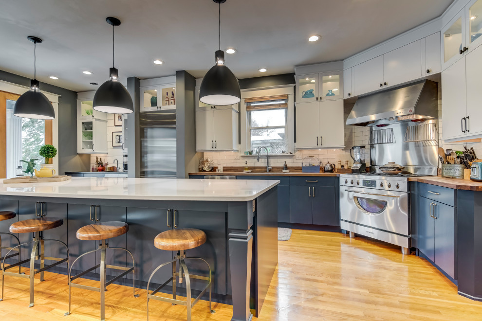 Bild på ett vintage brun brunt l-kök, med släta luckor, blå skåp, träbänkskiva, vitt stänkskydd, stänkskydd i tunnelbanekakel, rostfria vitvaror, mellanmörkt trägolv, en köksö och brunt golv
