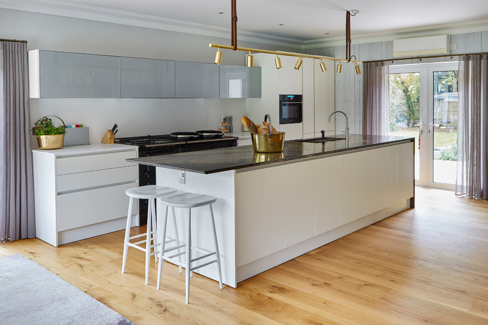 Inspiration for a contemporary galley open plan kitchen in Sussex with light hardwood flooring, an island, flat-panel cabinets, white cabinets, black appliances and brown floors.