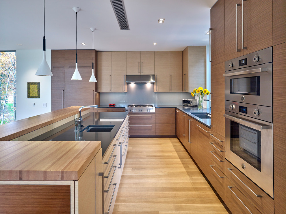Modern inredning av ett l-kök, med en undermonterad diskho, släta luckor, skåp i mellenmörkt trä, grått stänkskydd, rostfria vitvaror, mellanmörkt trägolv, en köksö och brunt golv