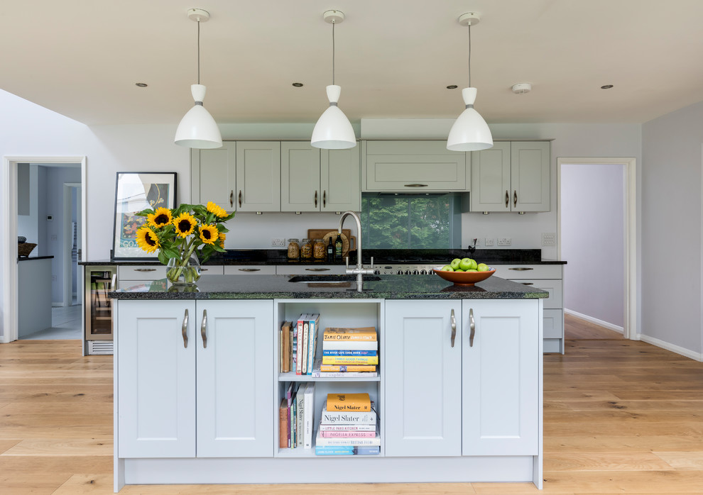 This is an example of a large classic single-wall open plan kitchen in Oxfordshire with a built-in sink, shaker cabinets, blue cabinets, granite worktops, blue splashback, glass sheet splashback, stainless steel appliances, light hardwood flooring and an island.
