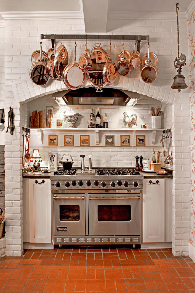 Foto på ett stort lantligt kök, med vita skåp, vitt stänkskydd, rostfria vitvaror och tegelgolv