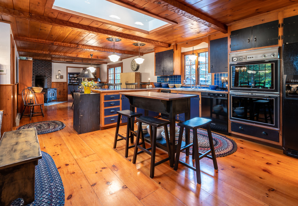 Cette photo montre une cuisine américaine nature en L avec un placard à porte plane, des portes de placard noires, un plan de travail en bois, une crédence bleue, un électroménager noir, un sol en bois brun, îlot, un sol orange et fenêtre au-dessus de l'évier.