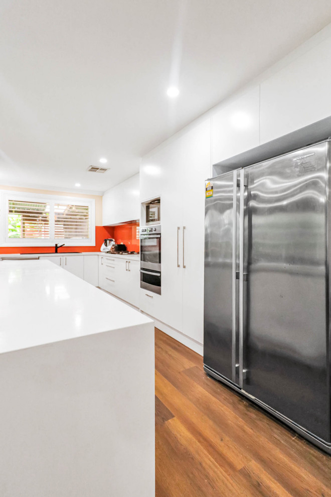 シドニーにあるお手頃価格の広いモダンスタイルのおしゃれなキッチン (ダブルシンク、フラットパネル扉のキャビネット、白いキャビネット、クオーツストーンカウンター、オレンジのキッチンパネル、ガラス板のキッチンパネル、シルバーの調理設備、クッションフロア、茶色い床、白いキッチンカウンター) の写真