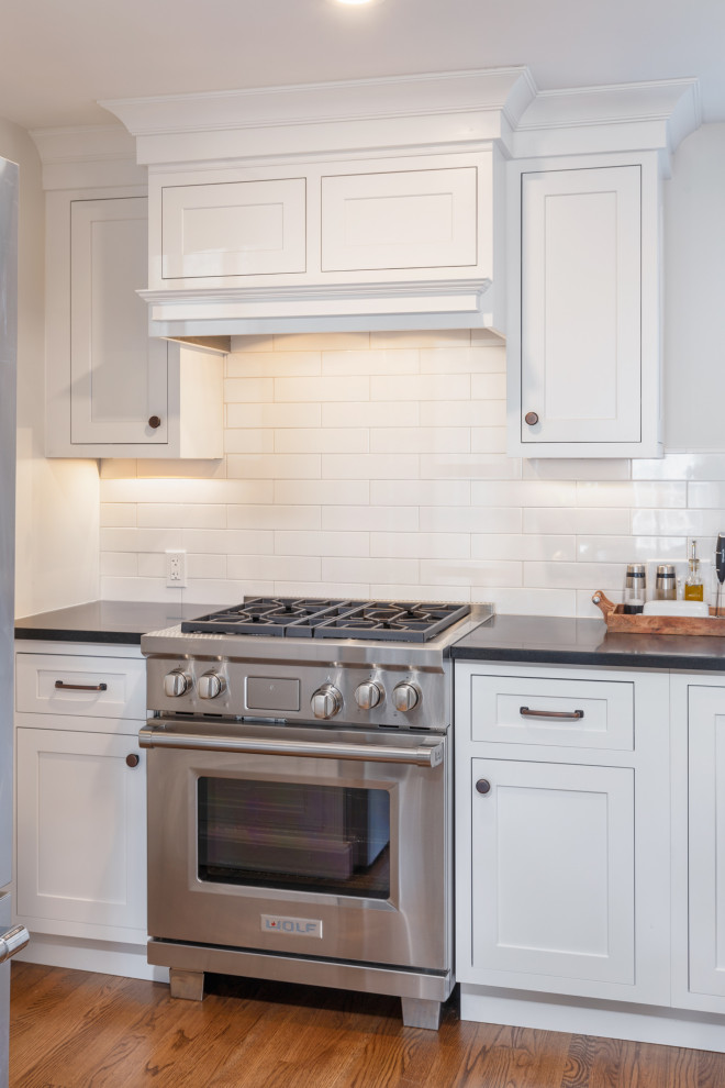ボストンにある高級な広いカントリー風のおしゃれなキッチン (エプロンフロントシンク、シェーカースタイル扉のキャビネット、白いキャビネット、クオーツストーンカウンター、白いキッチンパネル、サブウェイタイルのキッチンパネル、シルバーの調理設備、無垢フローリング、茶色い床、白いキッチンカウンター) の写真