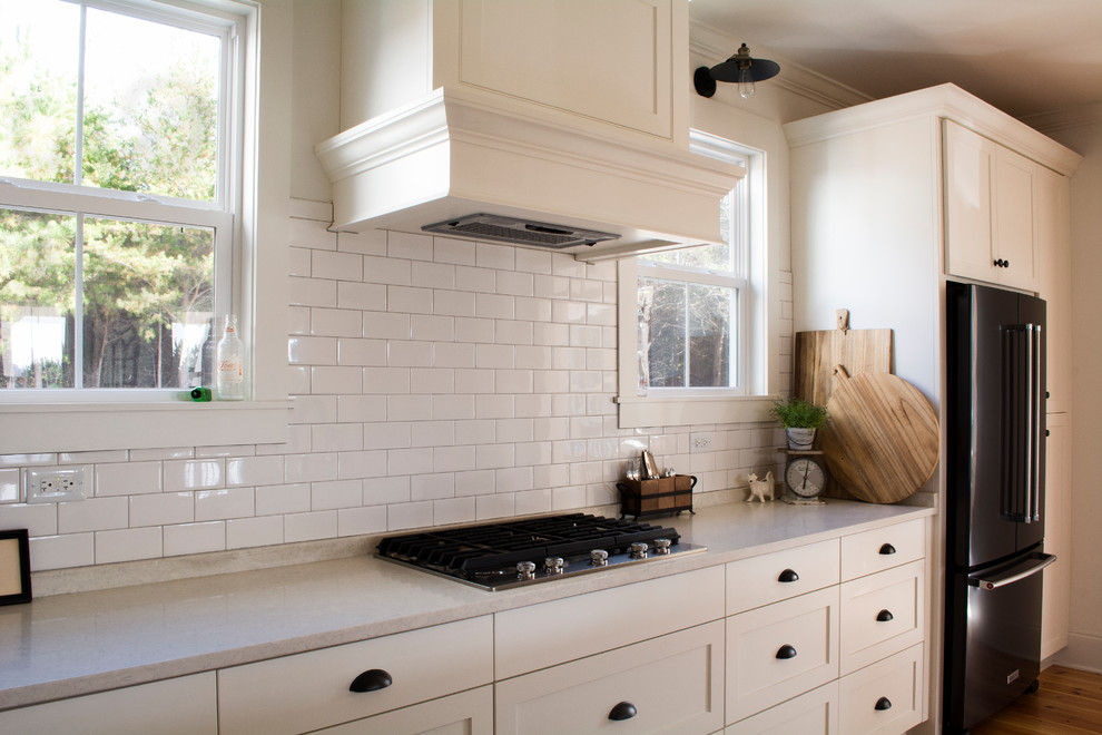 Foto på ett mellanstort lantligt vit l-kök, med en undermonterad diskho, skåp i shakerstil, vita skåp, bänkskiva i kvarts, vitt stänkskydd, stänkskydd i porslinskakel, svarta vitvaror, mellanmörkt trägolv, en köksö och brunt golv