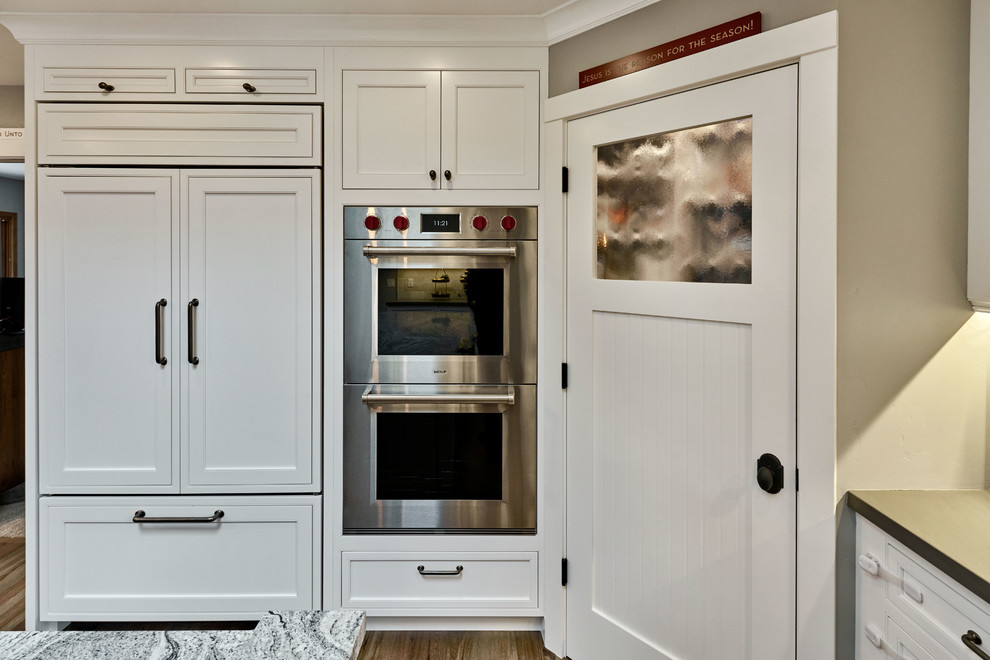 Foto på ett stort lantligt kök, med en rustik diskho, luckor med profilerade fronter, vita skåp, granitbänkskiva, grått stänkskydd, stänkskydd i stenkakel, integrerade vitvaror, ljust trägolv och en köksö