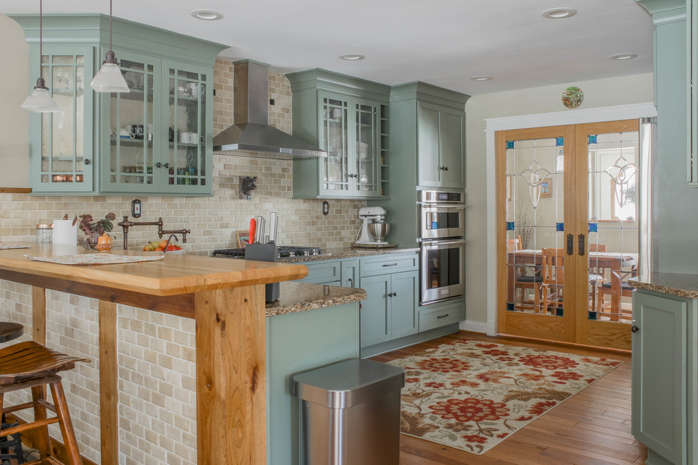 This is an example of a large farmhouse u-shaped kitchen/diner in Detroit with a belfast sink, shaker cabinets, green cabinets, engineered stone countertops, beige splashback, travertine splashback, stainless steel appliances, medium hardwood flooring, a breakfast bar, brown floors and brown worktops.