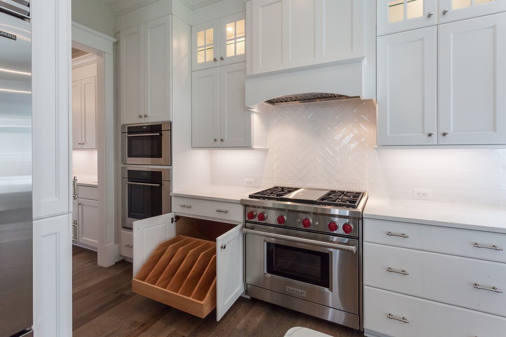 Photo of a rural l-shaped open plan kitchen in DC Metro with a belfast sink, recessed-panel cabinets, white cabinets, white splashback, metro tiled splashback, stainless steel appliances, medium hardwood flooring, an island, brown floors and white worktops.