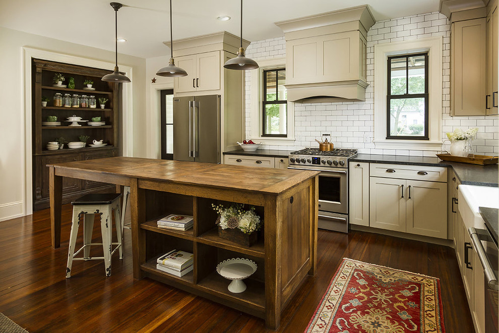 This is an example of a rural grey and cream l-shaped kitchen in DC Metro with a belfast sink, shaker cabinets, beige cabinets, white splashback, metro tiled splashback, stainless steel appliances, dark hardwood flooring, an island and brown floors.