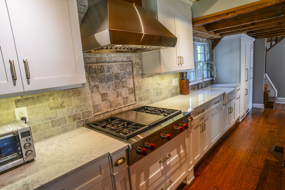 Example of a large farmhouse single-wall medium tone wood floor eat-in kitchen design in New York with a farmhouse sink, shaker cabinets, white cabinets, quartz countertops, beige backsplash, stone tile backsplash, stainless steel appliances and an island