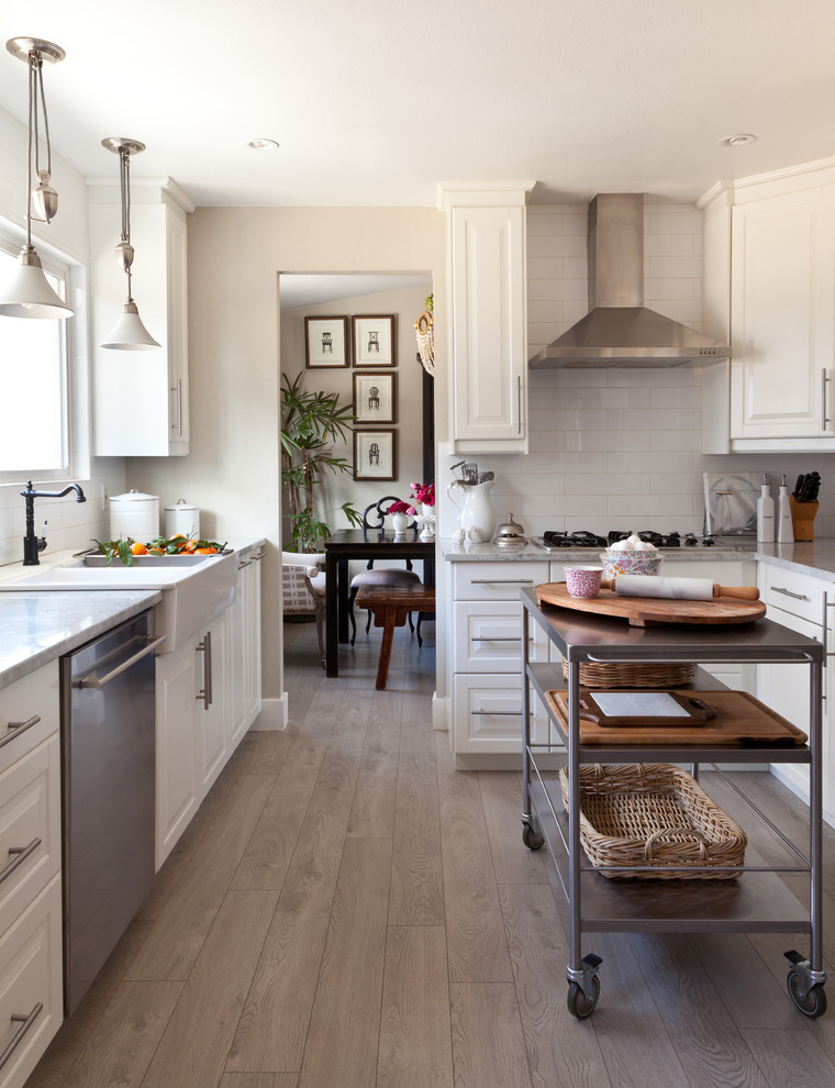 Bild på ett lantligt l-kök, med en rustik diskho, luckor med upphöjd panel, vita skåp, vitt stänkskydd, rostfria vitvaror, ljust trägolv och en köksö