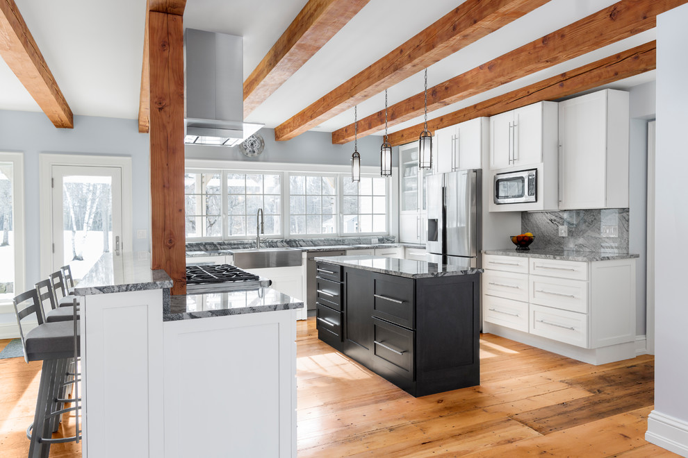 This is an example of a farmhouse u-shaped kitchen/diner in Portland Maine with a belfast sink, shaker cabinets, white cabinets, granite worktops, grey splashback, stone slab splashback, stainless steel appliances, medium hardwood flooring, an island, brown floors and grey worktops.