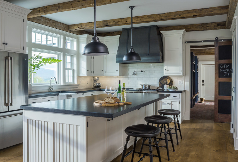 Mid-sized cottage u-shaped medium tone wood floor and brown floor kitchen photo in Other with a farmhouse sink, white cabinets, granite countertops, white backsplash, subway tile backsplash, stainless steel appliances, an island, black countertops and beaded inset cabinets