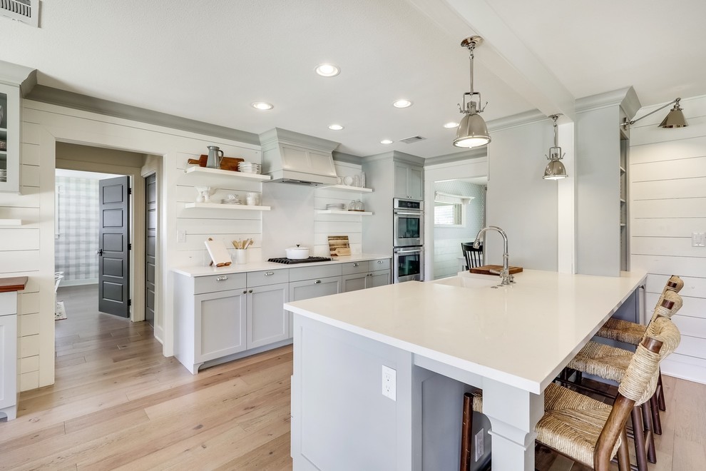 This is an example of a medium sized rural grey and cream galley kitchen/diner in Austin with a belfast sink, shaker cabinets, engineered stone countertops, light hardwood flooring, white worktops, grey cabinets, white splashback, stainless steel appliances, an island and beige floors.