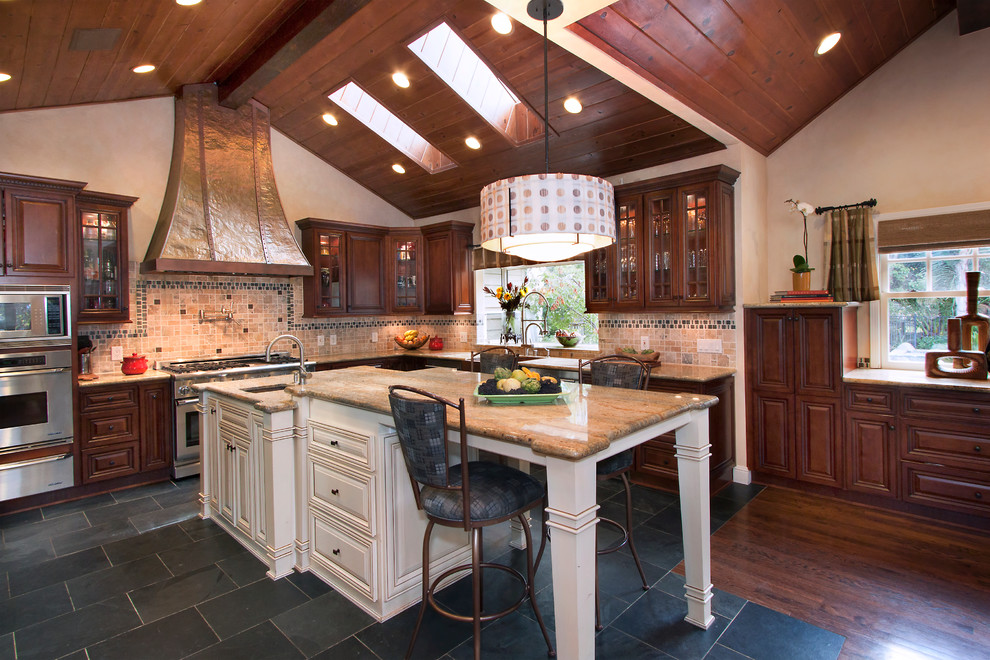 Exemple d'une grande cuisine américaine chic en L et bois foncé avec un évier de ferme, un placard avec porte à panneau surélevé, un plan de travail en granite, une crédence multicolore, une crédence en carrelage de pierre, un électroménager en acier inoxydable, un sol en travertin, îlot et un sol noir.