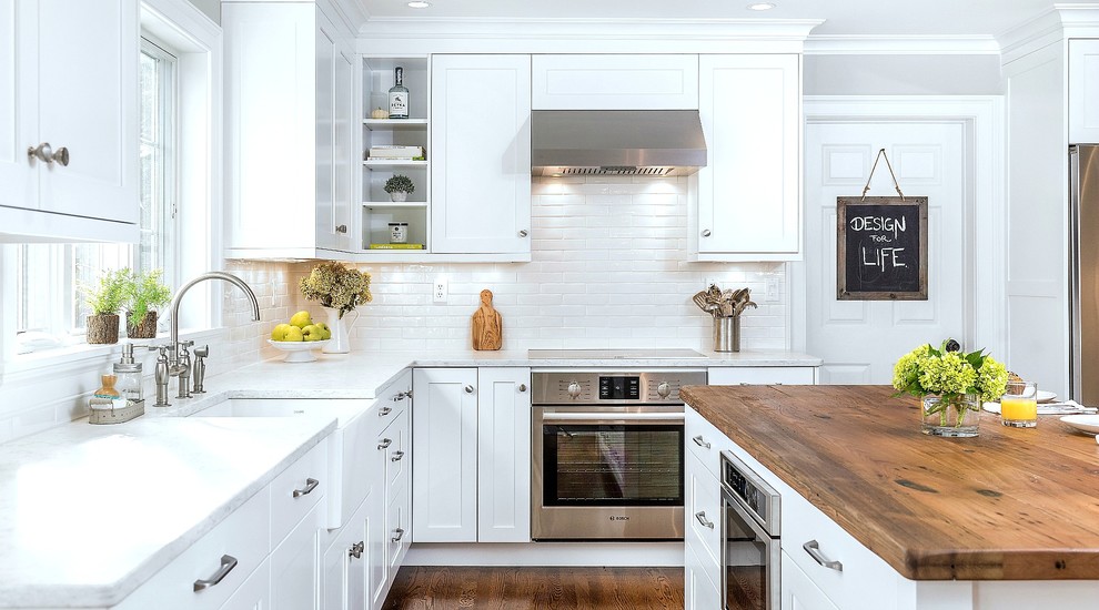 Enclosed kitchen - mid-sized transitional l-shaped medium tone wood floor and brown floor enclosed kitchen idea in Other with a farmhouse sink, recessed-panel cabinets, white cabinets, quartz countertops, white backsplash, ceramic backsplash, stainless steel appliances and an island