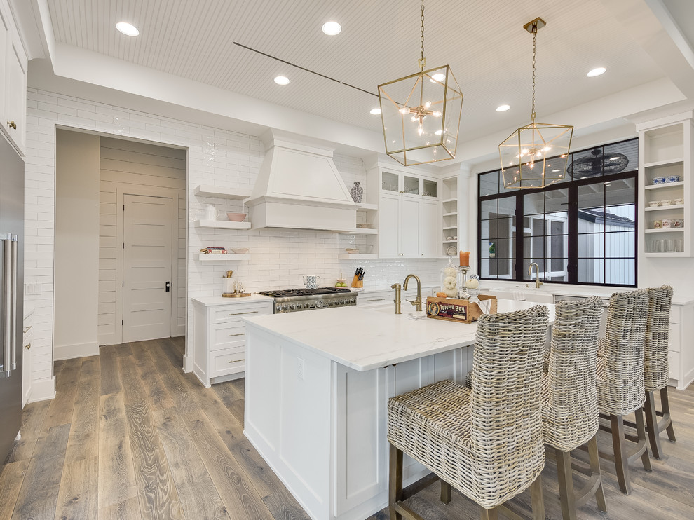 Photo of a large farmhouse u-shaped kitchen/diner in Austin with a belfast sink, recessed-panel cabinets, white cabinets, quartz worktops, white splashback, metro tiled splashback, stainless steel appliances, light hardwood flooring, an island, grey floors and white worktops.