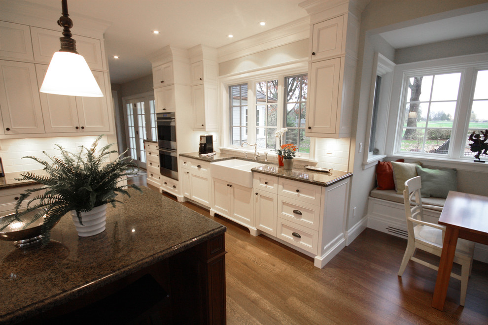 Photo of a large classic u-shaped open plan kitchen in Toronto with a belfast sink, recessed-panel cabinets, white cabinets, granite worktops, white splashback, ceramic splashback, stainless steel appliances, medium hardwood flooring and an island.