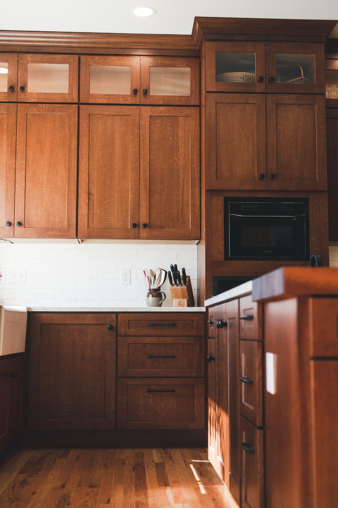シアトルにある高級な中くらいなカントリー風のおしゃれなキッチン (エプロンフロントシンク、落し込みパネル扉のキャビネット、中間色木目調キャビネット、珪岩カウンター、白いキッチンパネル、サブウェイタイルのキッチンパネル、黒い調理設備、無垢フローリング、茶色い床、白いキッチンカウンター) の写真