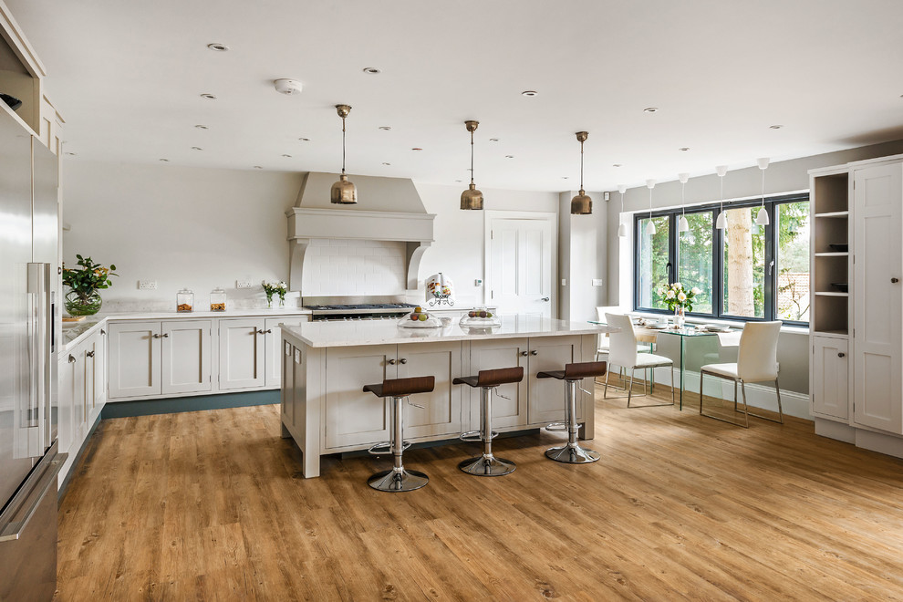 Klassische Wohnküche in L-Form mit Schrankfronten im Shaker-Stil, weißen Schränken, Küchenrückwand in Weiß, Rückwand aus Metrofliesen, Küchengeräten aus Edelstahl, hellem Holzboden und Kücheninsel in Surrey