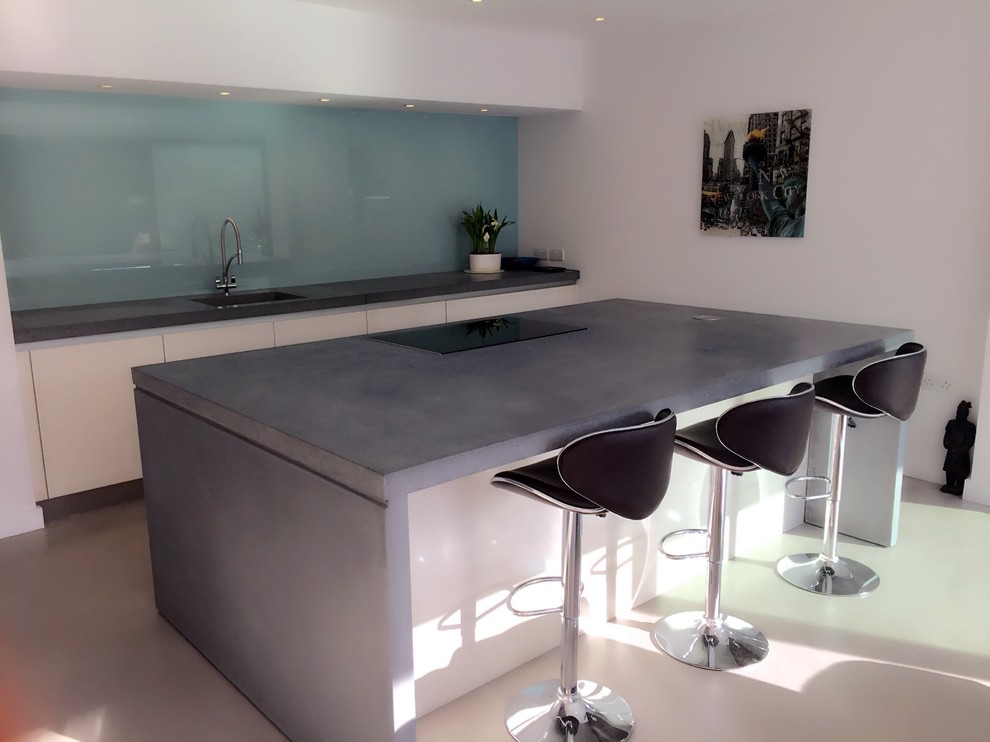 Offene Moderne Küche mit Einbauwaschbecken, weißen Schränken, Betonarbeitsplatte, Küchenrückwand in Blau, Glasrückwand, Elektrogeräten mit Frontblende, Betonboden und Kücheninsel in Surrey