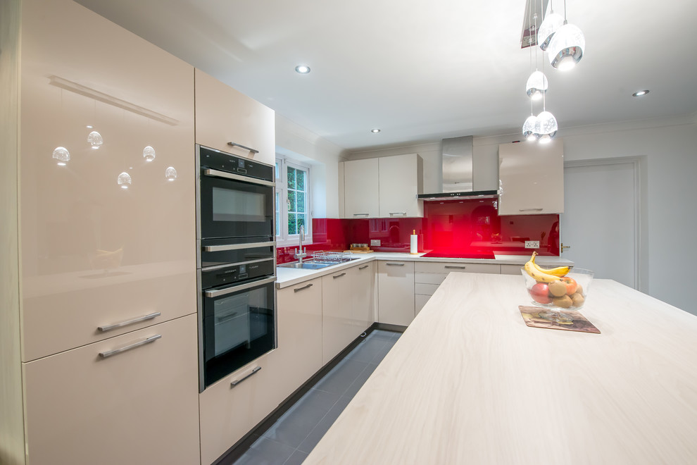 This is an example of a contemporary l-shaped kitchen in London with a double-bowl sink, flat-panel cabinets, light wood cabinets, onyx worktops, red splashback, glass sheet splashback, black appliances and an island.