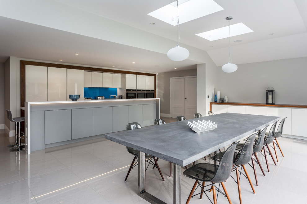 This is an example of an expansive contemporary grey and white single-wall kitchen/diner in Hertfordshire with a double-bowl sink, flat-panel cabinets, white cabinets, composite countertops, blue splashback, glass sheet splashback, integrated appliances, porcelain flooring, an island, grey floors, white worktops and feature lighting.
