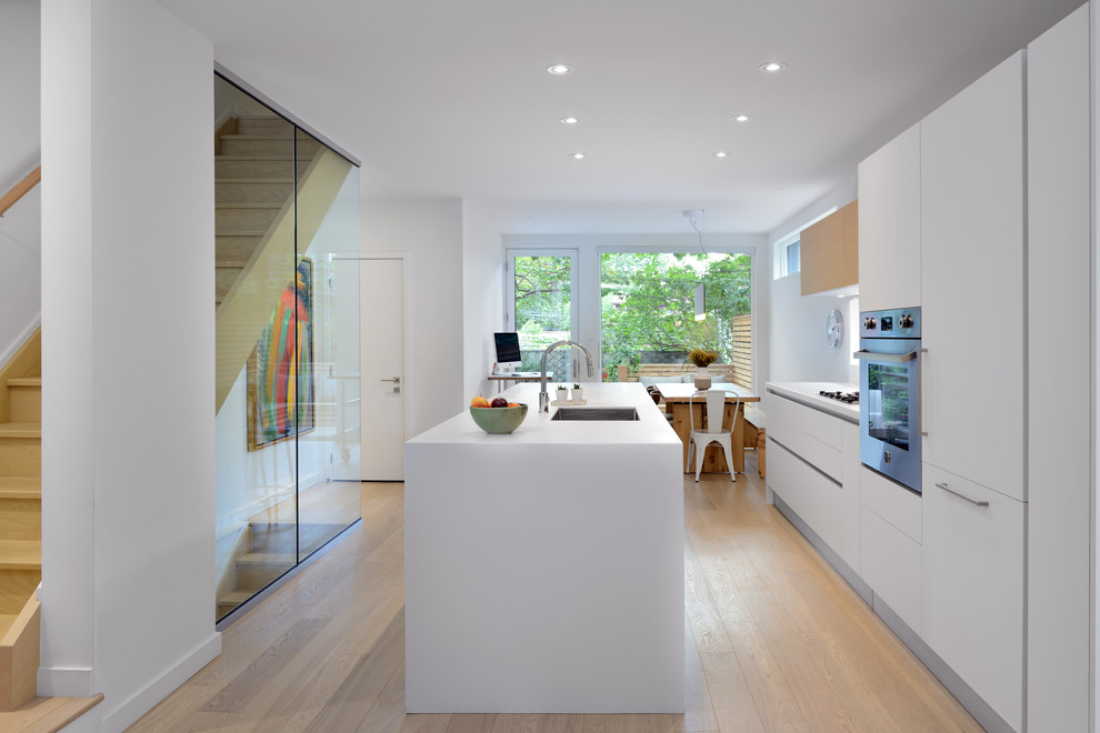 Idéer för ett modernt kök och matrum, med en enkel diskho, släta luckor, vita skåp, rostfria vitvaror, ljust trägolv och en köksö
