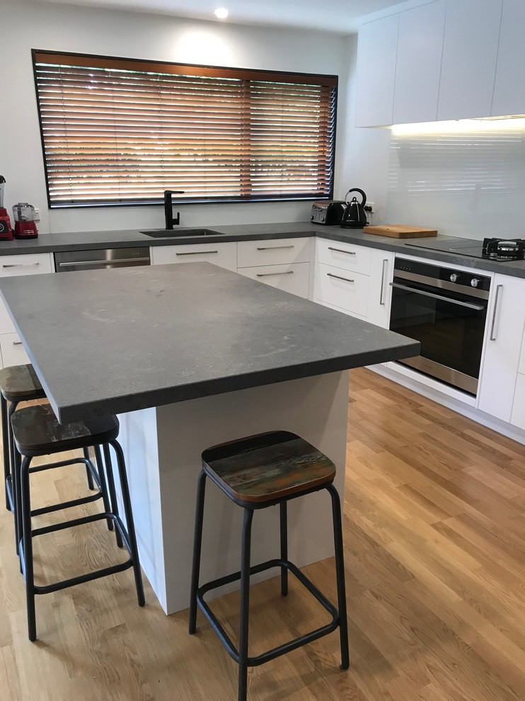 This is an example of a large contemporary l-shaped open plan kitchen in Christchurch with a single-bowl sink, flat-panel cabinets, white cabinets, engineered stone countertops, white splashback, glass sheet splashback, stainless steel appliances, medium hardwood flooring, an island and brown floors.