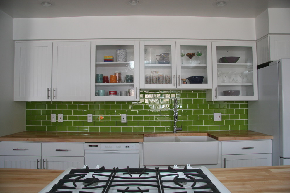 Photo of a medium sized country single-wall kitchen in Phoenix with a belfast sink, glass-front cabinets, white cabinets, wood worktops, green splashback, metro tiled splashback, white appliances, dark hardwood flooring and an island.