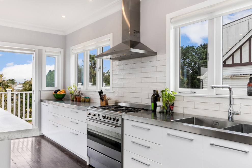 Example of a mid-sized trendy galley dark wood floor and brown floor eat-in kitchen design in Auckland with a double-bowl sink, white cabinets, stainless steel countertops, white backsplash, subway tile backsplash, stainless steel appliances and no island