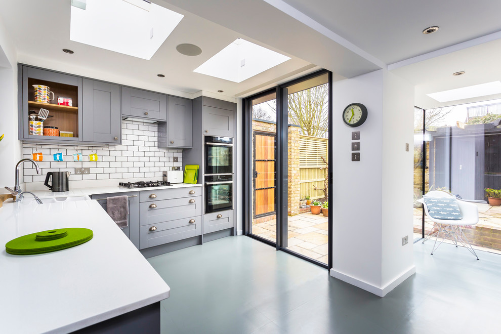 Inspiration for a small contemporary l-shaped kitchen/diner in Surrey with a belfast sink, shaker cabinets, grey cabinets, composite countertops, white splashback, ceramic splashback, black appliances, vinyl flooring, a breakfast bar, grey floors and white worktops.