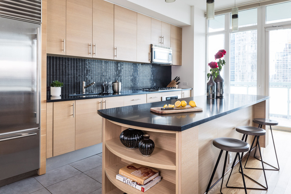 Trendy single-wall gray floor kitchen photo in New York with an undermount sink, flat-panel cabinets, light wood cabinets, black backsplash, stainless steel appliances, an island and black countertops