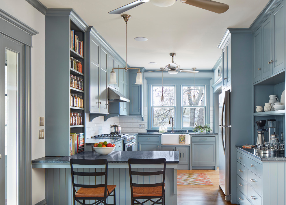 Foto på ett amerikanskt grå u-kök, med en rustik diskho, skåp i shakerstil, blå skåp, vitt stänkskydd, stänkskydd i tunnelbanekakel, rostfria vitvaror, ljust trägolv och en halv köksö