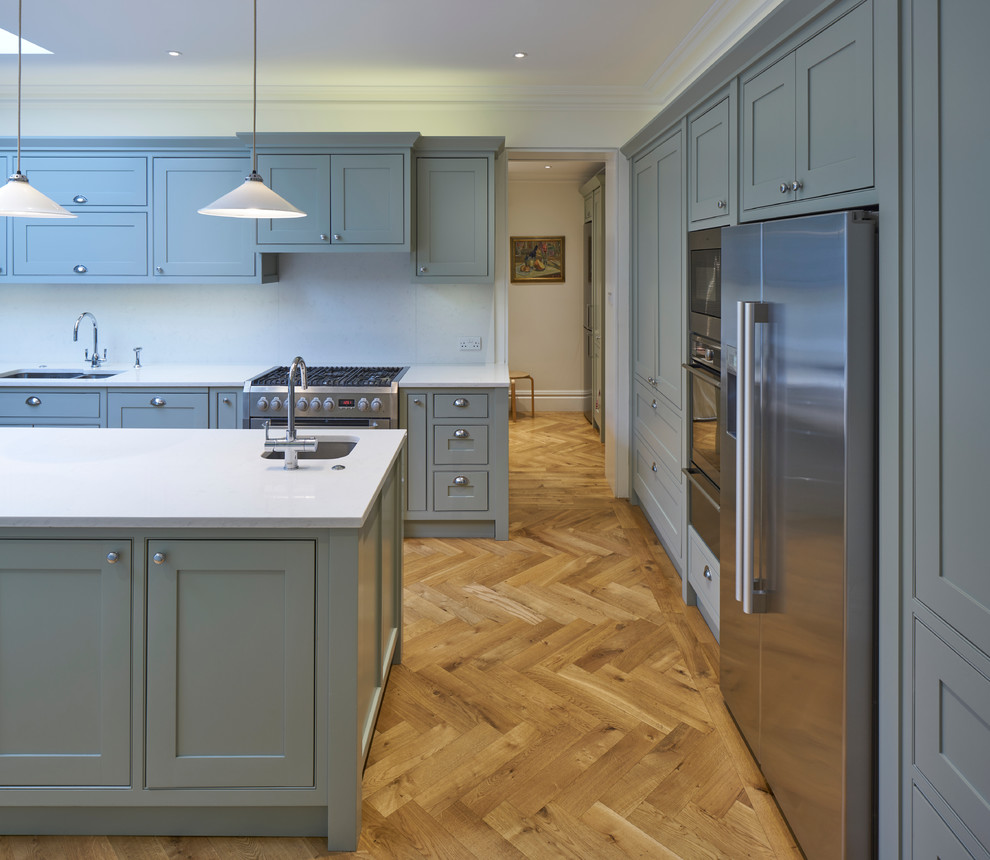 Design ideas for a large traditional l-shaped open plan kitchen in London with a double-bowl sink, recessed-panel cabinets, grey cabinets, composite countertops, white splashback, stainless steel appliances, medium hardwood flooring and multiple islands.