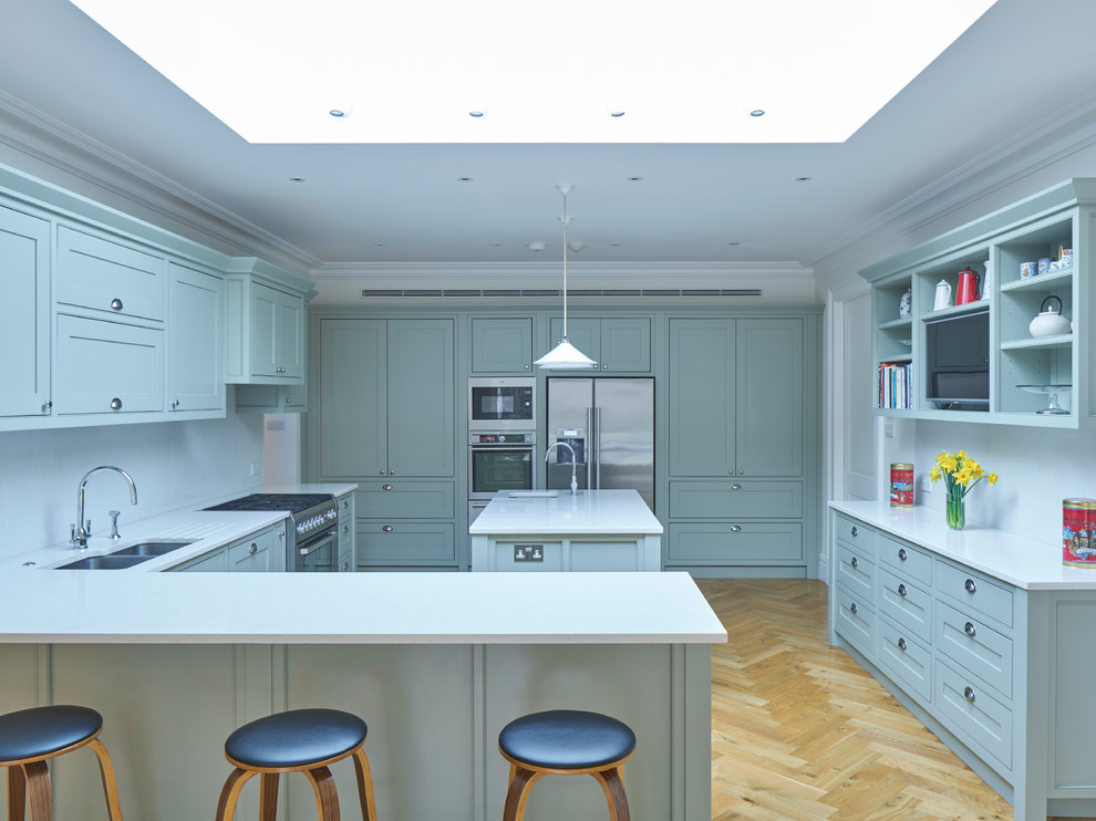 Large traditional l-shaped open plan kitchen in London with a double-bowl sink, recessed-panel cabinets, grey cabinets, composite countertops, white splashback, stainless steel appliances, medium hardwood flooring and multiple islands.