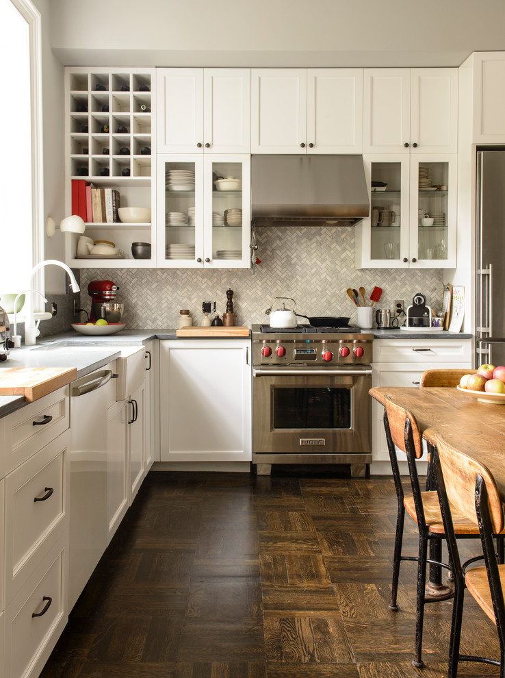 Example of a classic l-shaped dark wood floor and brown floor eat-in kitchen design in New York with white cabinets, a farmhouse sink, shaker cabinets, gray backsplash, stainless steel appliances and no island