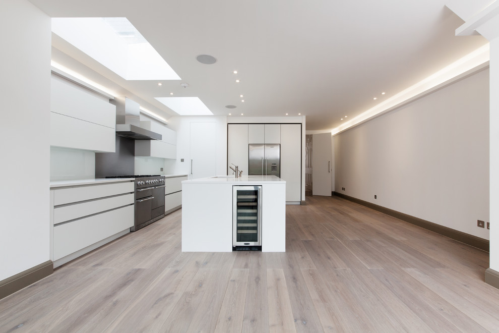 Medium sized contemporary l-shaped open plan kitchen in London with a submerged sink, flat-panel cabinets, white cabinets, quartz worktops, white splashback, glass tiled splashback, stainless steel appliances, light hardwood flooring and an island.