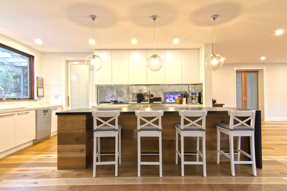 Modern inredning av ett stort kök, med en undermonterad diskho, släta luckor, vita skåp, bänkskiva i kvarts, stänkskydd med metallisk yta, spegel som stänkskydd, rostfria vitvaror, mellanmörkt trägolv och en köksö