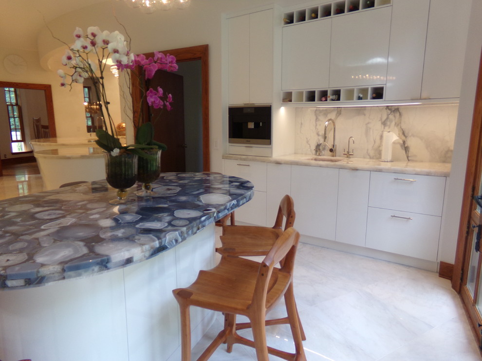 Expansive contemporary u-shaped kitchen pantry in New York with a belfast sink, flat-panel cabinets, white cabinets, engineered stone countertops, white splashback, stone slab splashback, stainless steel appliances, porcelain flooring and an island.